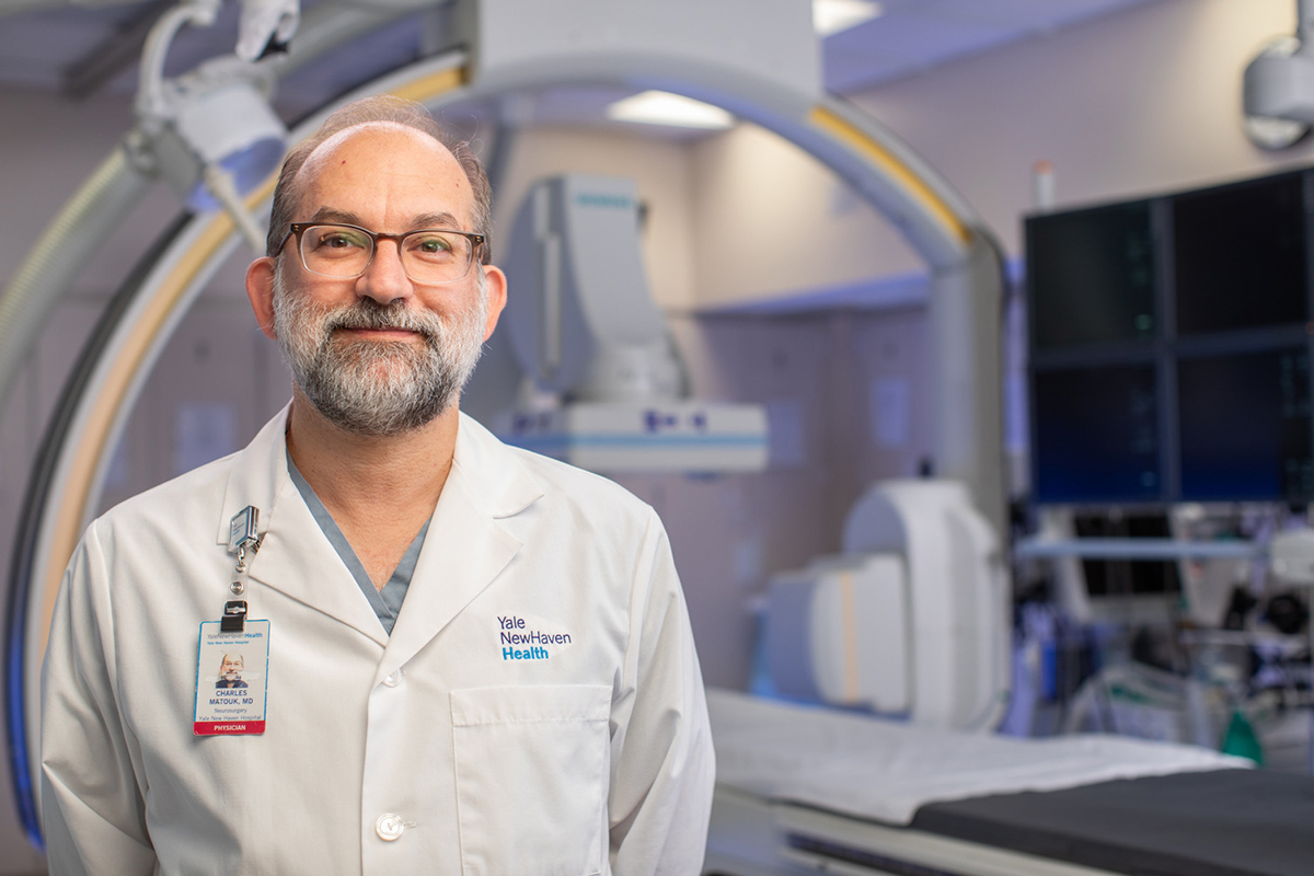 A balding man with a beard, glasses and a white coat, poses in an interior setting in front of large medical equipment.