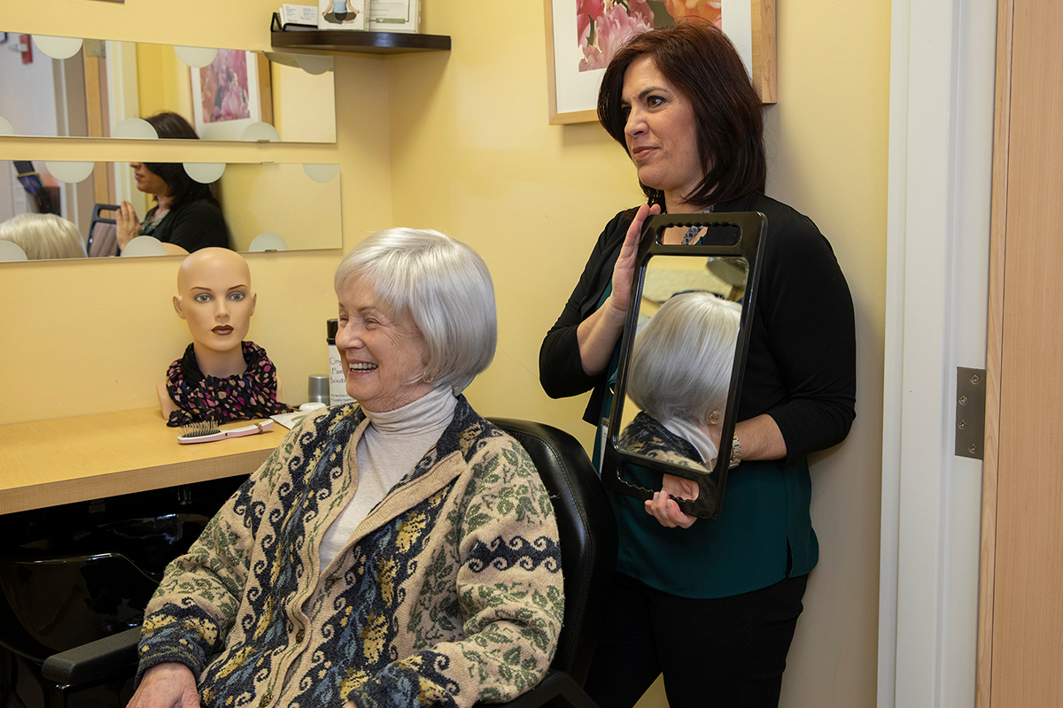 A woman wearing a gray wig, seated and looking to the left at an off-screen mirror, smiles as a female hairdresser stands behind her with a hand mirror.